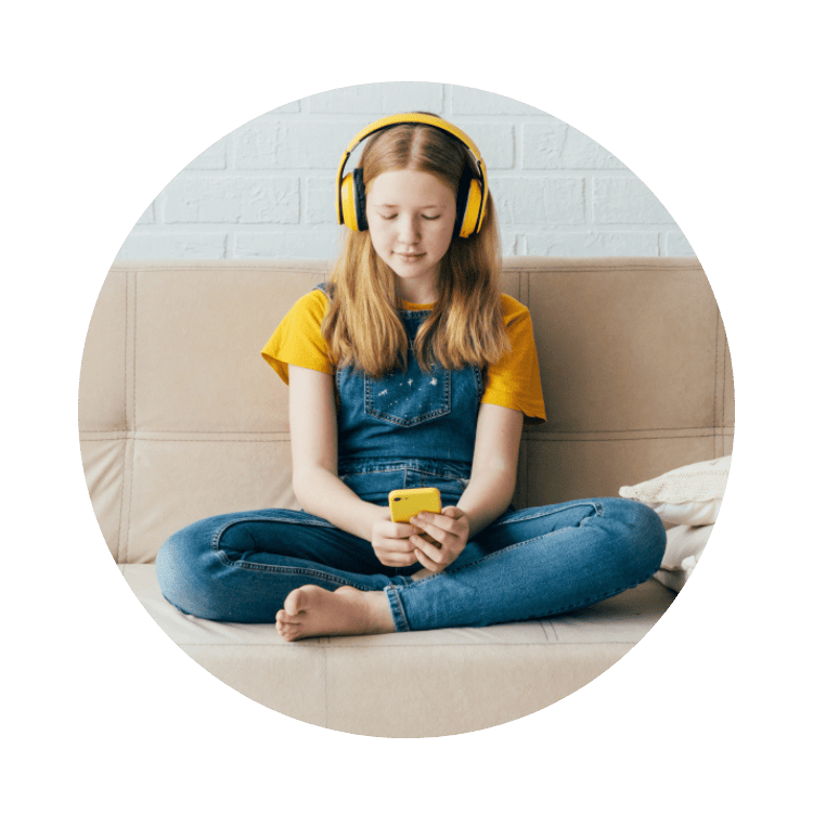 eatappie - Ein junges Mädchen in Jeanslatzhose und gelbem T-Shirt sitzt im Schneidersitz auf einem Sofa, trägt gelbe Kopfhörer und blickt auf ihr Smartphone. Sie benutzt dabei die „eatappie“-App, die speziell für Jugendliche mit Magersucht oder Bulimie entwickelt wurde.